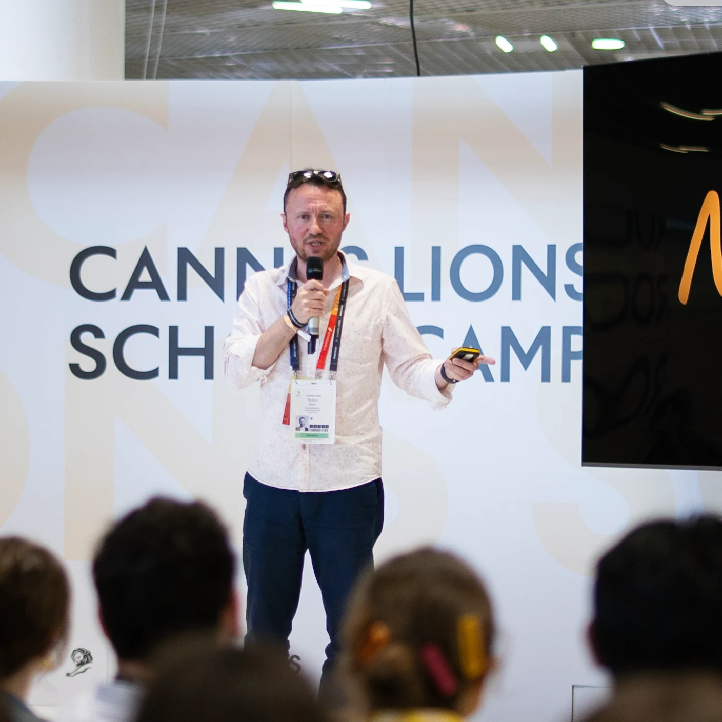 Man wearing a light patterned shirt and dark pants speaking into a microphone while holding a device, standing in front of a Cannes Lions School backdrop with an audience in the foreground.