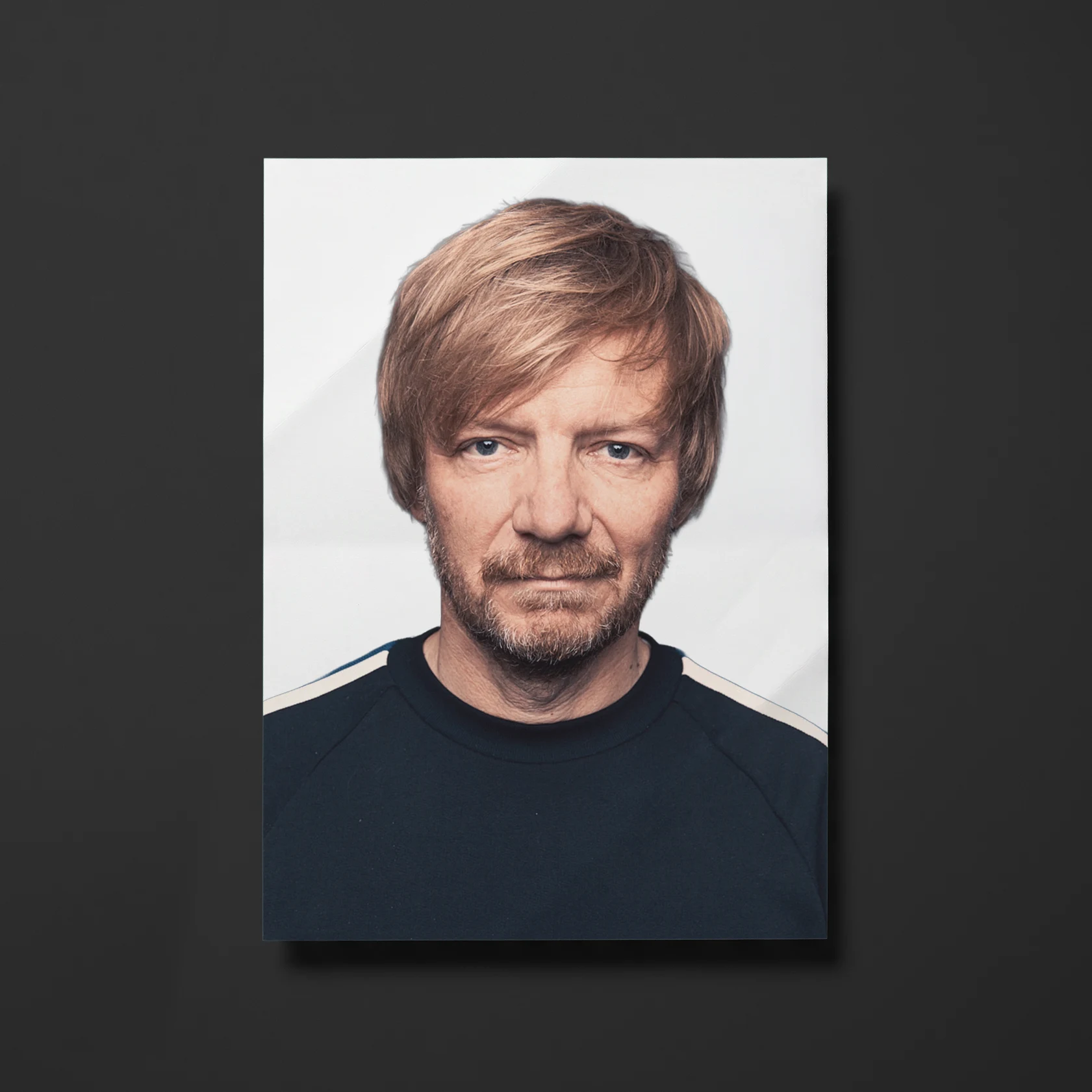 Portrait of a man with light brown hair and a short beard, wearing a dark blue shirt with light stripes on the shoulders, looking directly at the camera with a neutral expression on a plain white background.
