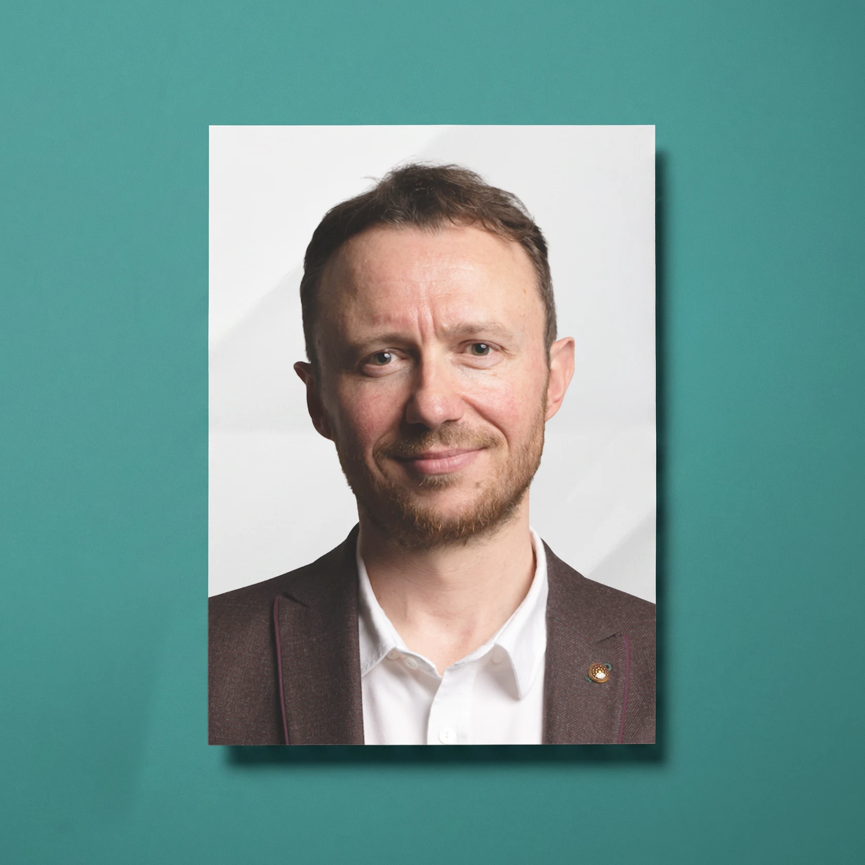 Headshot of a smiling man with short brown hair and beard wearing a white shirt and dark blazer with a pin on the lapel, against a plain white background.