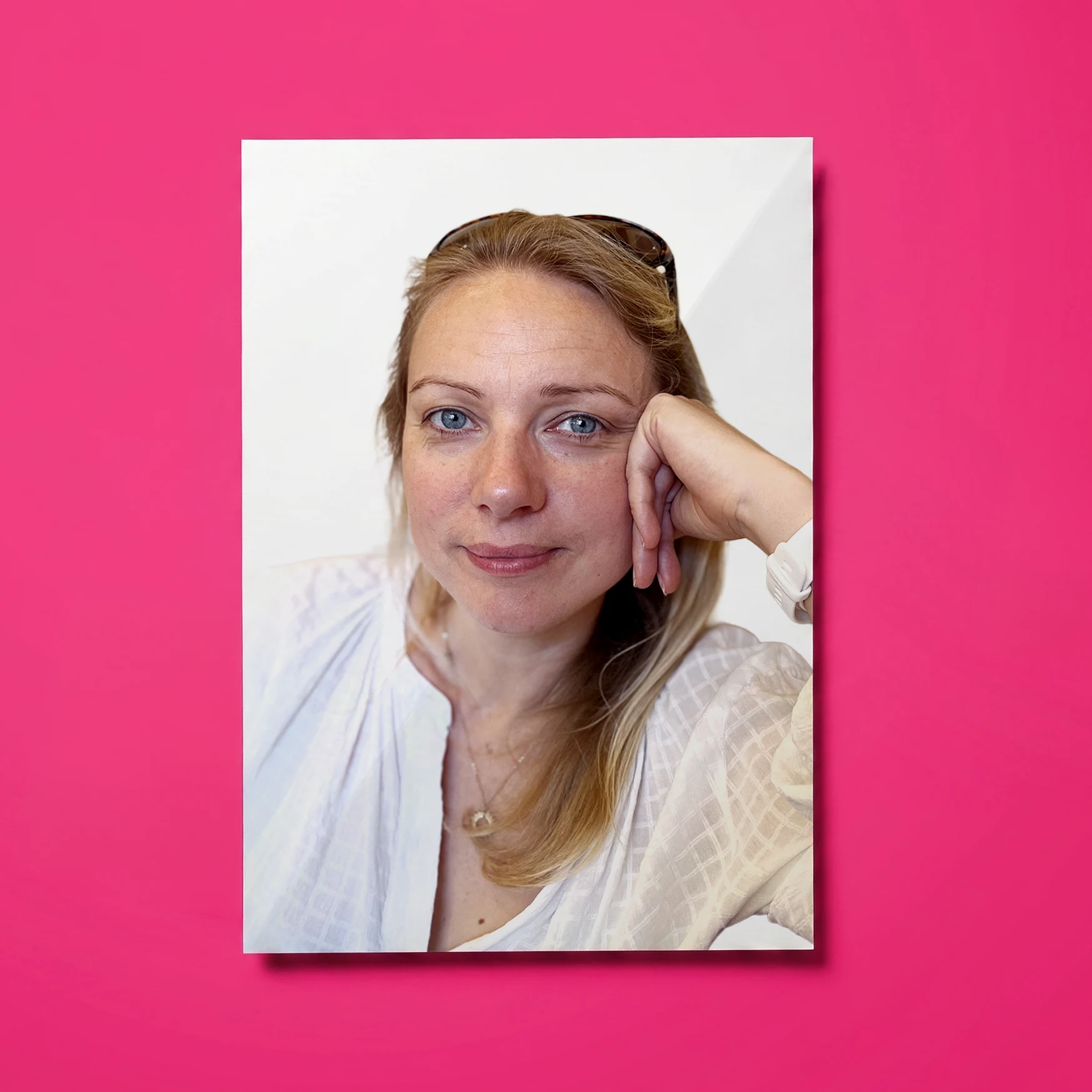 Portrait of a woman with blonde hair and blue eyes resting her head on her hand, wearing a white blouse and a watch, against a white background with a pink border.