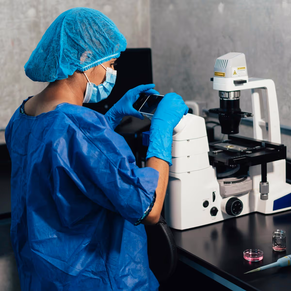 Scientist wearing blue protective gear and gloves using a smartphone to capture an image of a microscope in a lab.