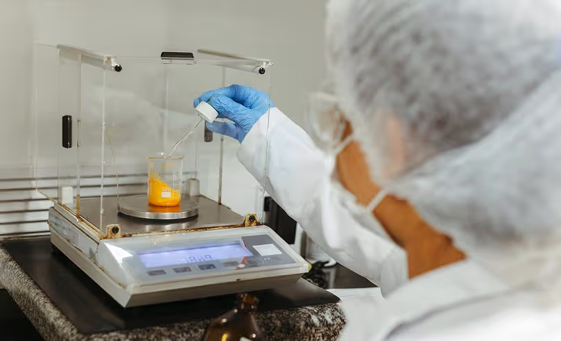 Scientist in protective gear weighing orange liquid in a beaker on a precision scale in a laboratory.