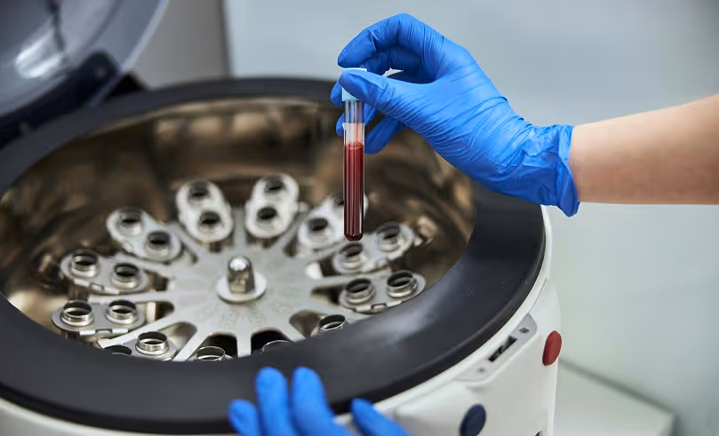Gloved hands holding a test tube with blood above a centrifuge machine.