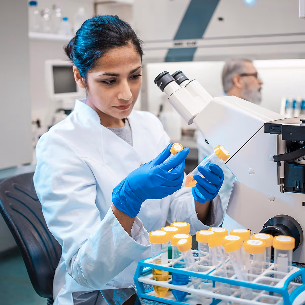 Female scientist in blue gloves examining a test tube next to a microscope in a laboratory.