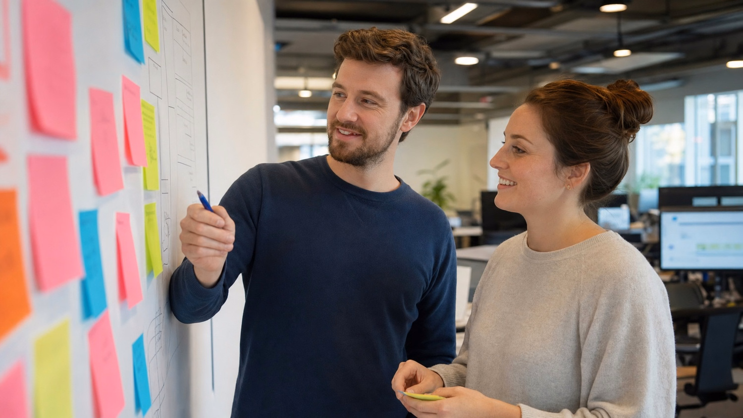 Two Caucasian individuals in their 30s stand at a wall covered in colorful stick