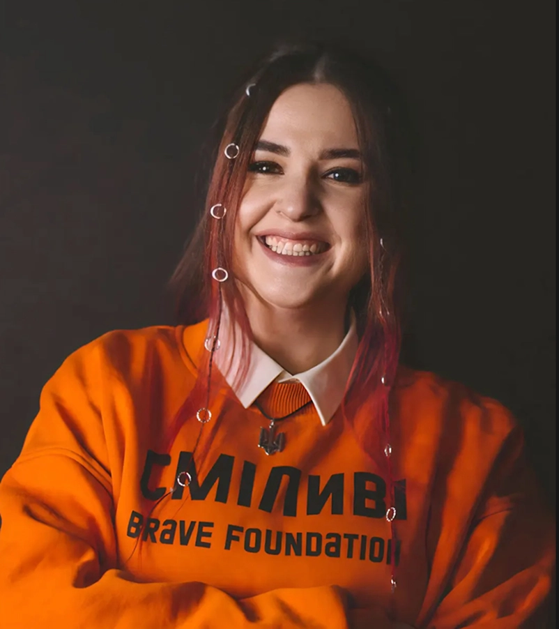 Smiling woman with red hair wearing an orange sweatshirt that says 'Brave Foundation' and a white collared shirt underneath.