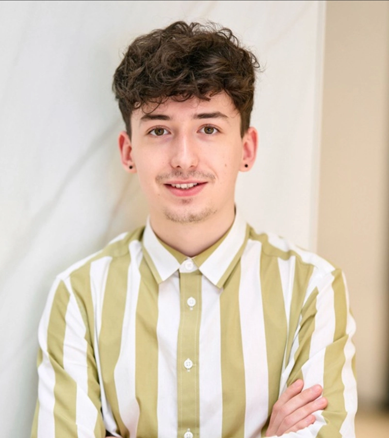 Young man with curly dark hair, wearing a green and white vertical striped shirt, smiling with arms crossed.
