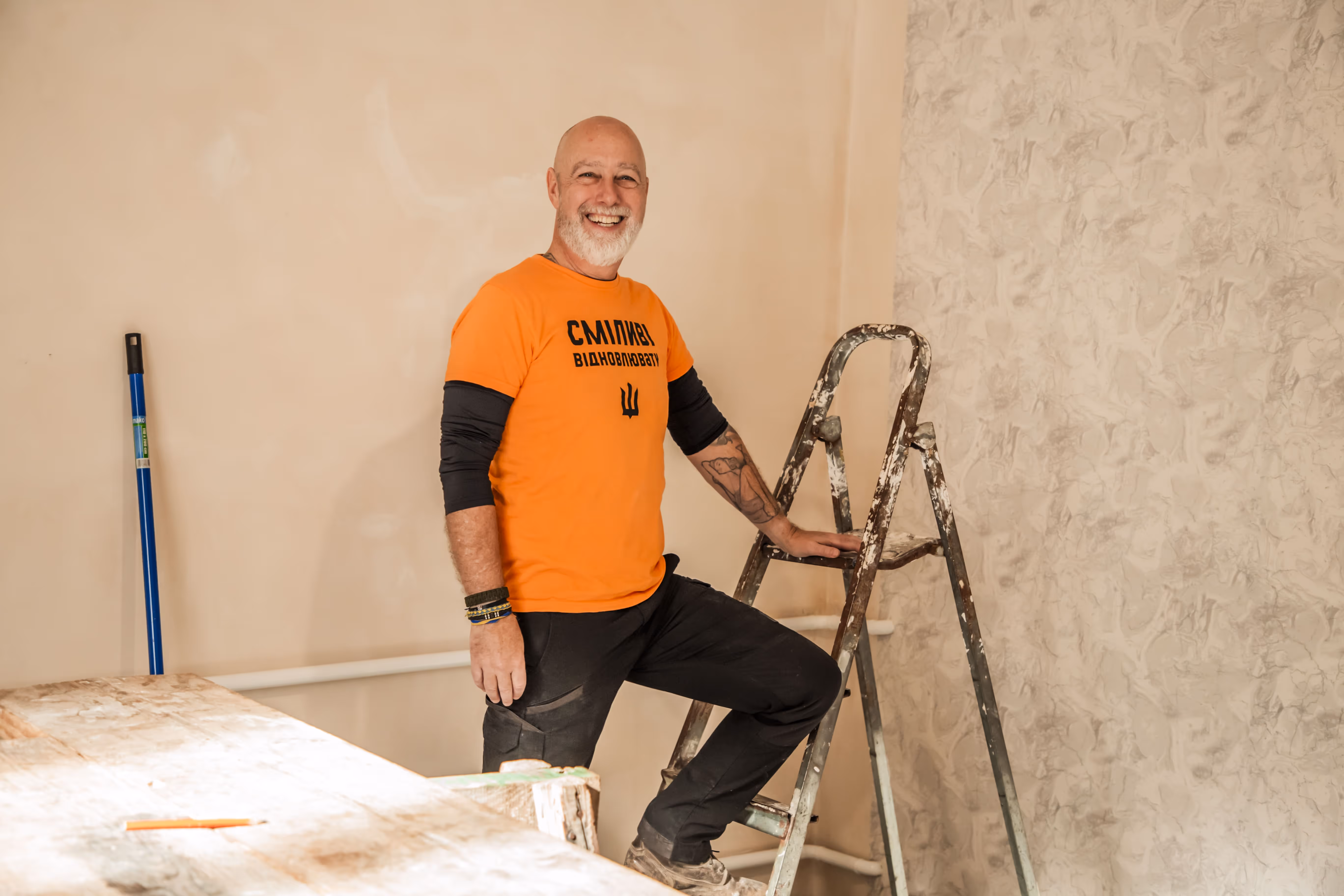 Smiling man in an orange shirt and black pants standing on a paint-spattered ladder inside a room under renovation.