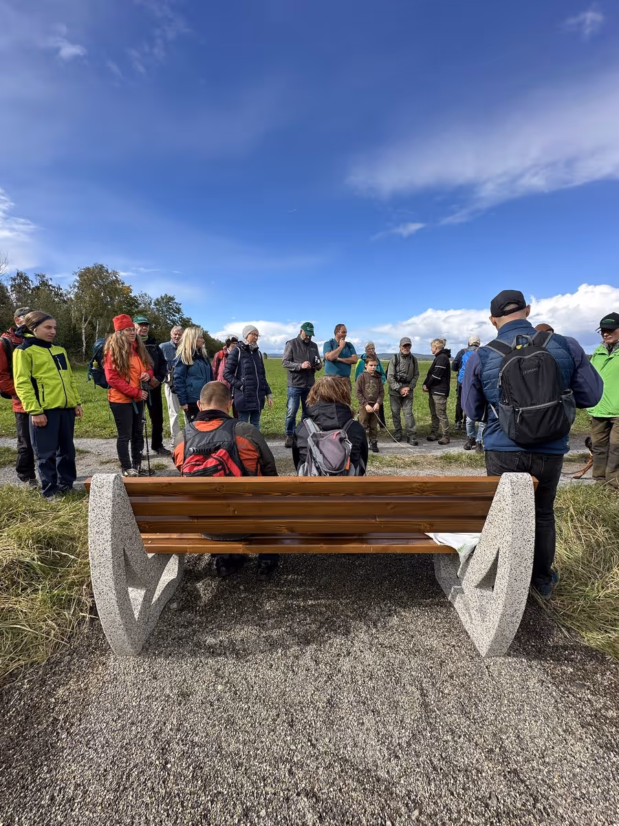 Sitzbank von hinten. Zwei Personen sitzen darauf. Mehere stehen davor. Es handelt sich um eine Gruppe Wandernder.