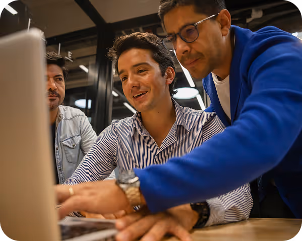 Three men collaborating at a wooden table in a modern office. One man is pointing at a laptop screen while another looks on, all engaged in discussion.
