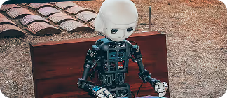 A humanoid robot sitting on a wooden bench, with a white head and mechanical body. It is positioned outdoors, surrounded by grass and tiles in the background.