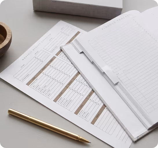 Open planner with finance tracker and budget sheets, gold pen on gray desk surface nearby.