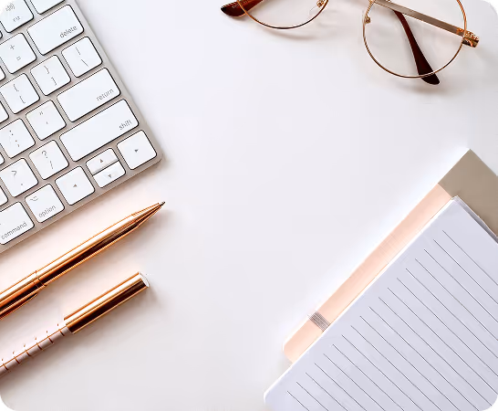 Rose gold pens, keyboard, glasses, and lined notepad on white desk from overhead view.