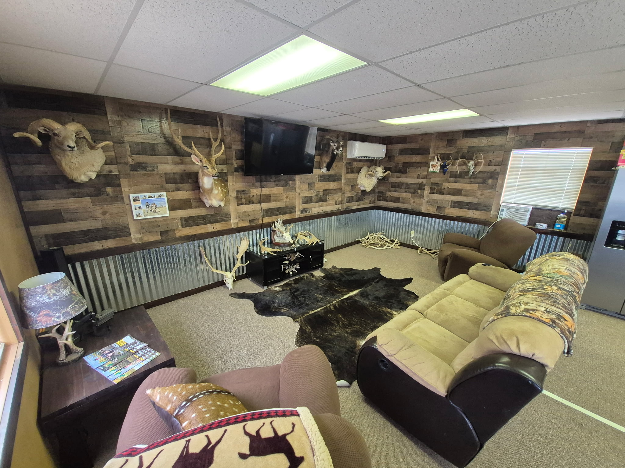 Cozy rustic room with animal head mounts, antlers, brown chairs, sofa, black rug, and TV on wood-paneled wall.