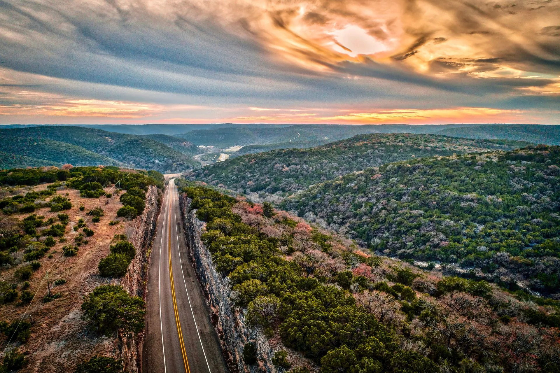A road cutting through hilly terrain covered with dense green and autumn-colored trees under a dramatic cloudy sunset sky.