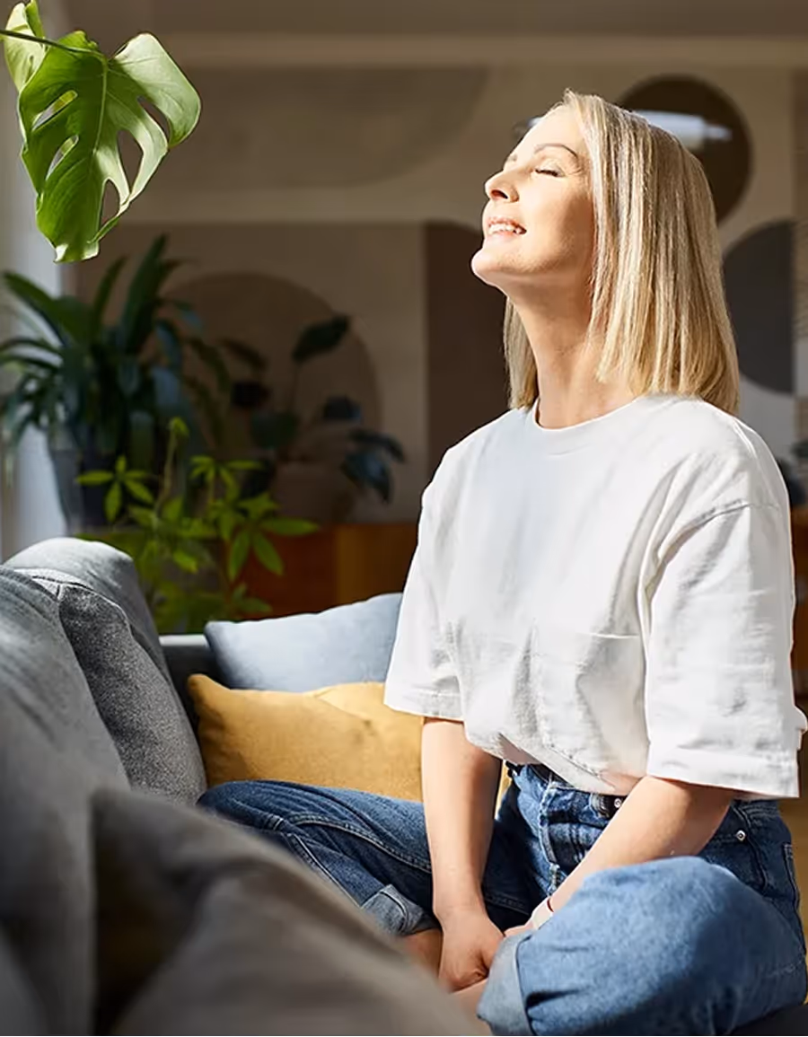 Mujer sonriente con camiseta blanca y jeans relajándose en un sofá junto a plantas verdes.