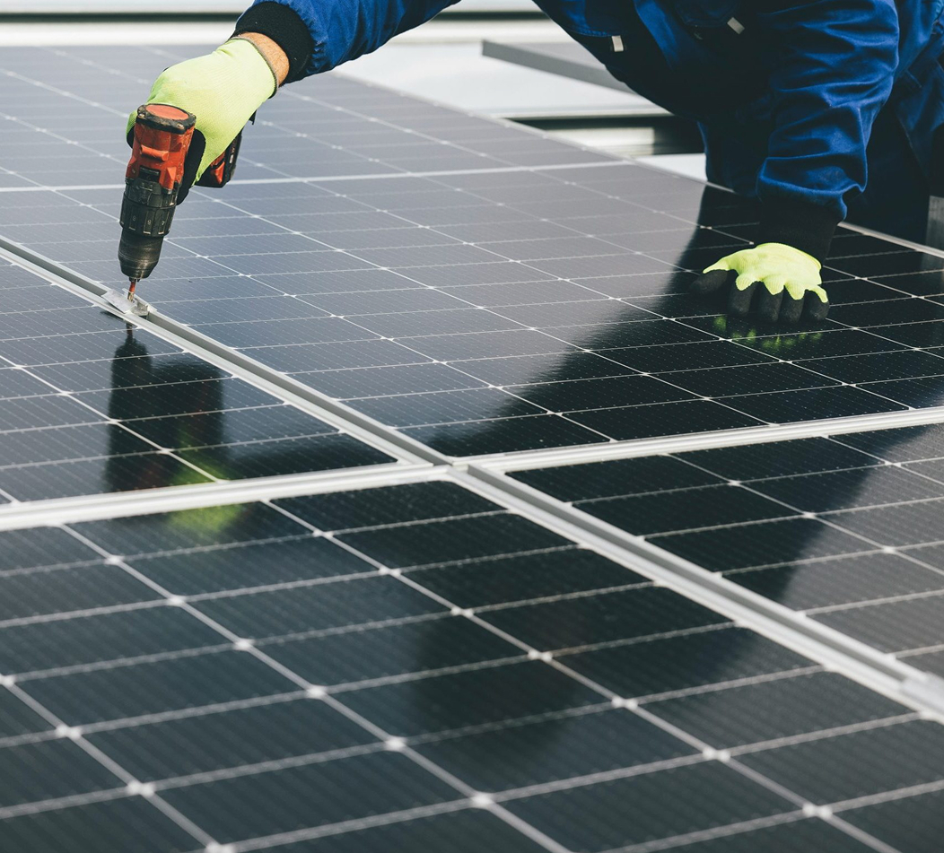 Trabajador instalando paneles solares usando un taladro y guantes amarillos fluorescentes.
