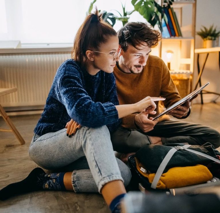 Pareja sentada en el suelo mirando una tablet mientras preparan una maleta.