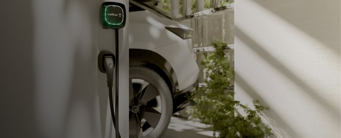 Coche eléctrico blanco cargándose en un cargador de pared Wallbox en un garaje con luz natural.