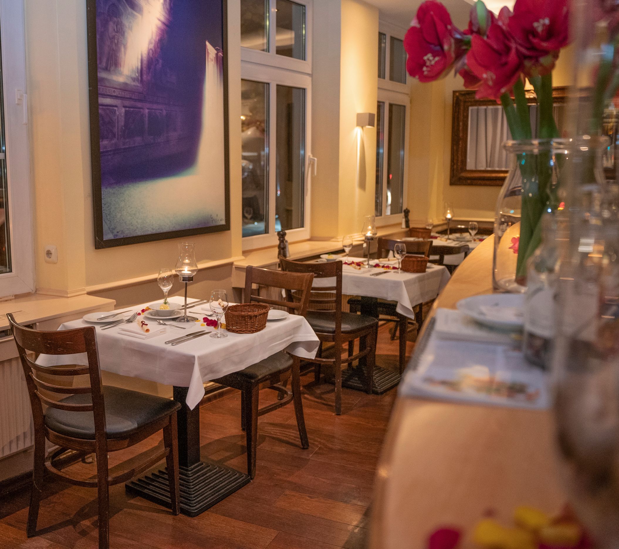 Gemütliches Restaurant mit gedeckten Tischen, weißen Tischdecken, Holzstühlen und einem großen Gemälde an der Wand.