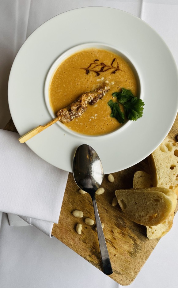 Schale mit cremiger Erdnusssuppe, garniert mit einem mit Erdnusssauce überzogenen Fleischspieß, frischen Korianderblättern und einem Deko-Muster aus dunkler Soße, daneben ein Löffel und Scheiben Weißbrot auf einem Holzbrett.