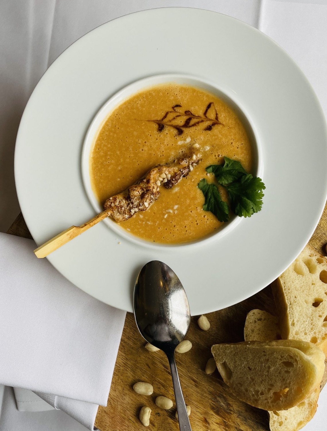 Cremesuppe mit einem Fleischspieß, Korianderblättern und gerösteten Nüssen auf einem weißen Teller, daneben ein Löffel und mehrere Scheiben Brot auf einem Holzbrett.