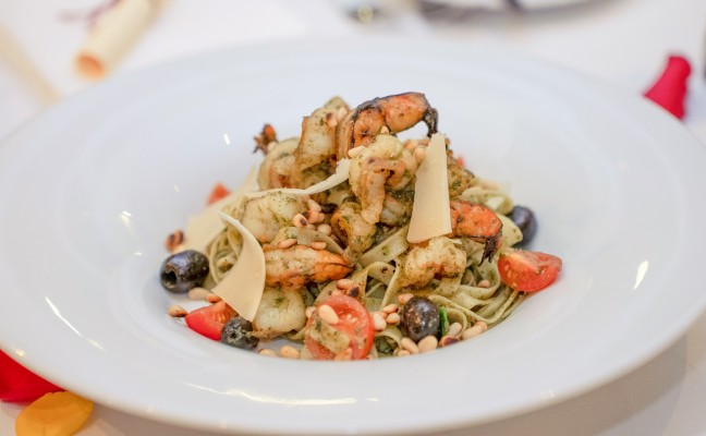 Teller mit Pasta, Garnelen, Kirschtomaten, schwarzen Oliven, Pinienkernen und Parmesanspänen.