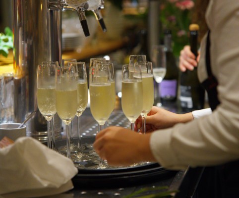 Mehrere mit Sekt gefüllte Champagnergläser auf einem Tablett, eine Person hält eines der Gläser in der Hand.