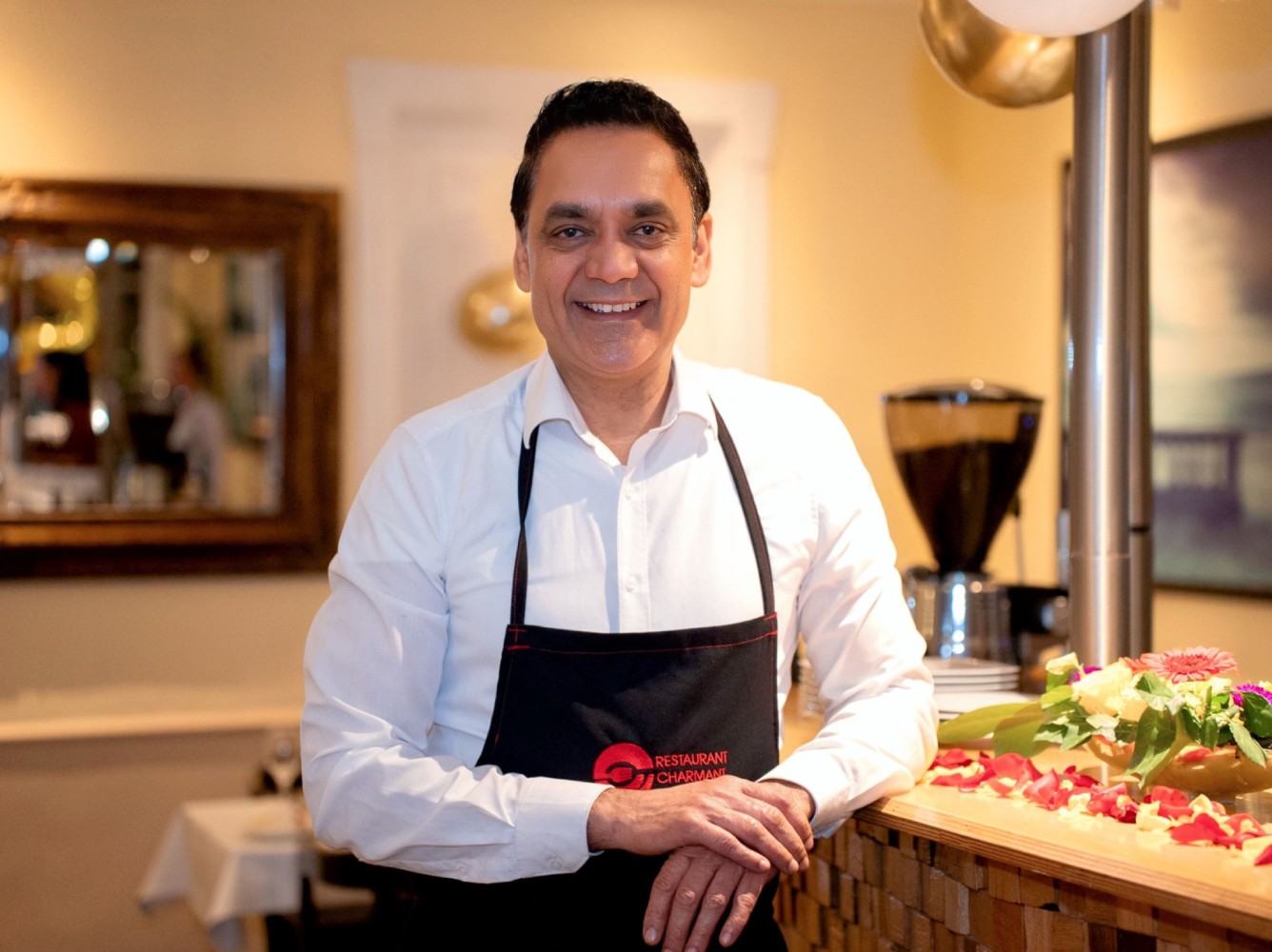 Der Besitzer vom Restaurant Charmant Dalip Singh in weißem Hemd und schwarzer Schürze lehnt an der Bar eines Restaurants mit Blumen und Rosenblättern.