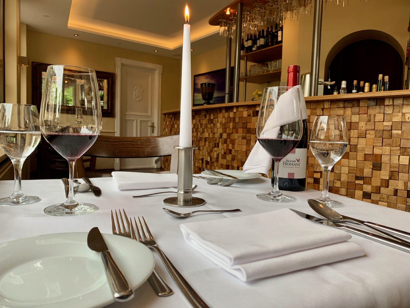 Weiß gedeckter Tisch in einem Restaurant mit Weingläsern, rotem Wein, Kerze und einer Weinflasche im Hintergrund.
