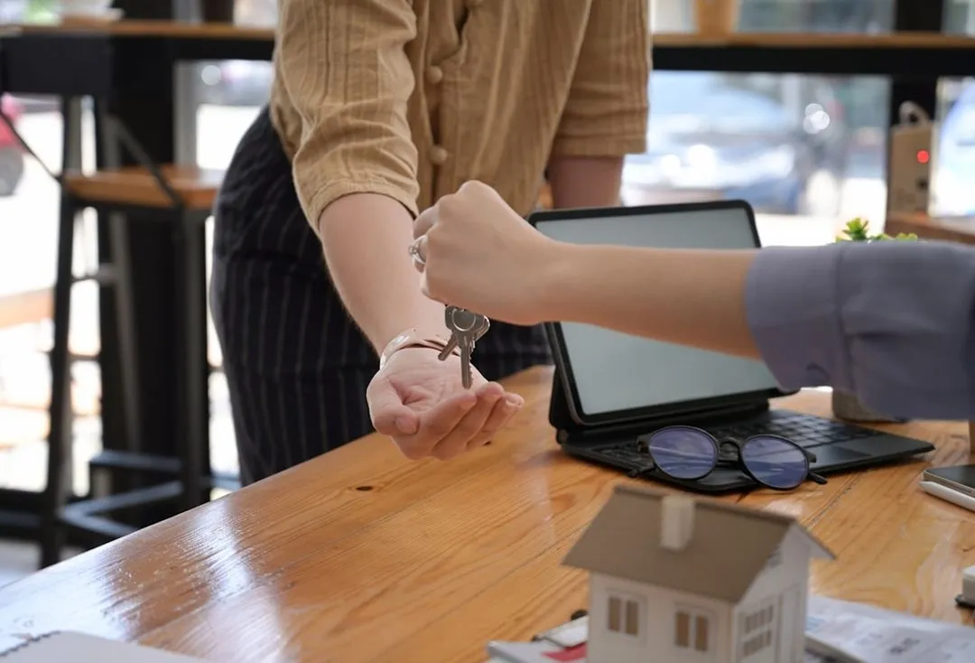 Remise de clés entre deux personnes à un bureau, avec un modèle de maison et un ordinateur portable en arrière-plan.