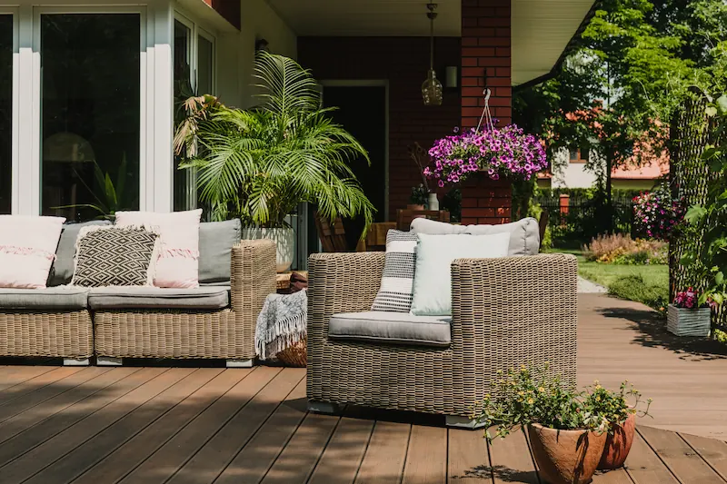 Wicker armchair and sofa with cushions on a wooden terrace with plants and hanging flowers.