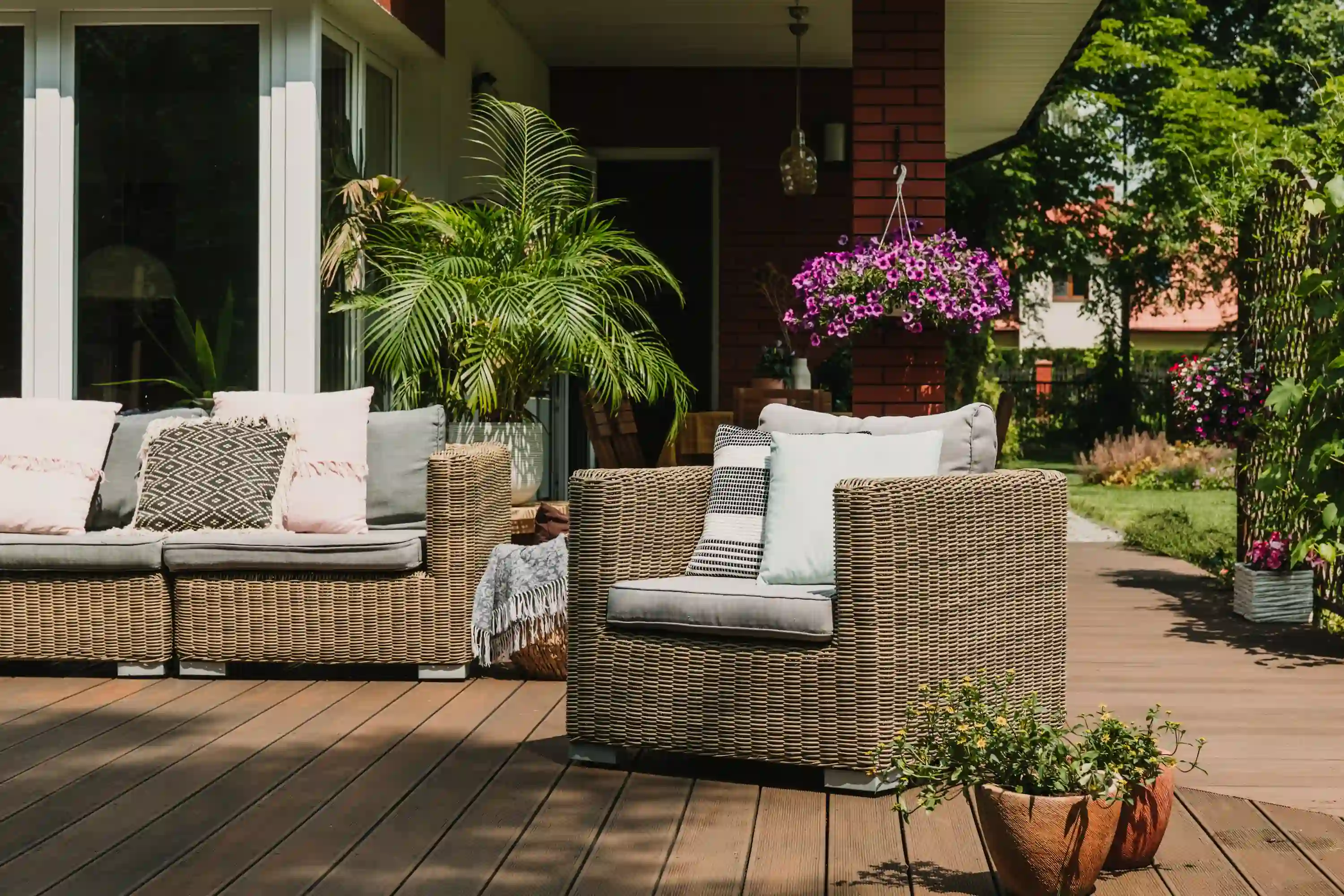Wicker armchair and sofa with cushions on a wooden patio next to potted plants and hanging flowers.