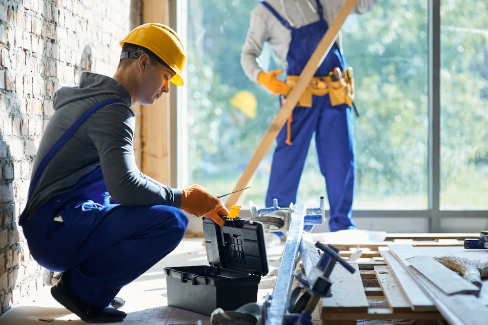 Construction workers in helmets and gloves working with tools and wood inside a building under renovation.