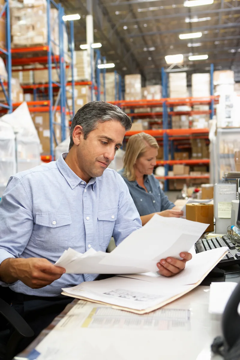 Two business colleagues reviewing paperwork at a desk in a warehouse with shelves of boxes.