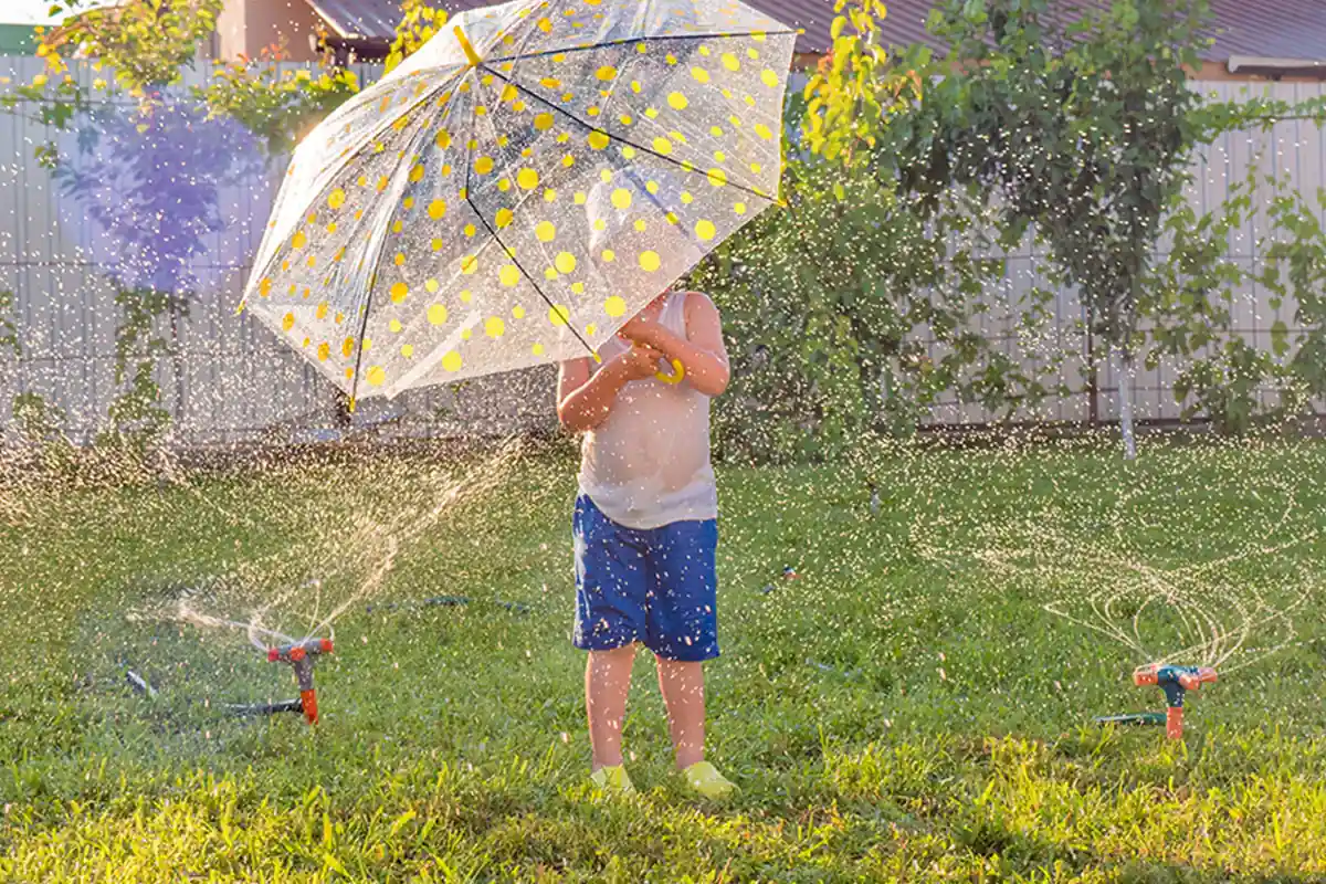 child holding umbrella amidst sprinklers in backyard