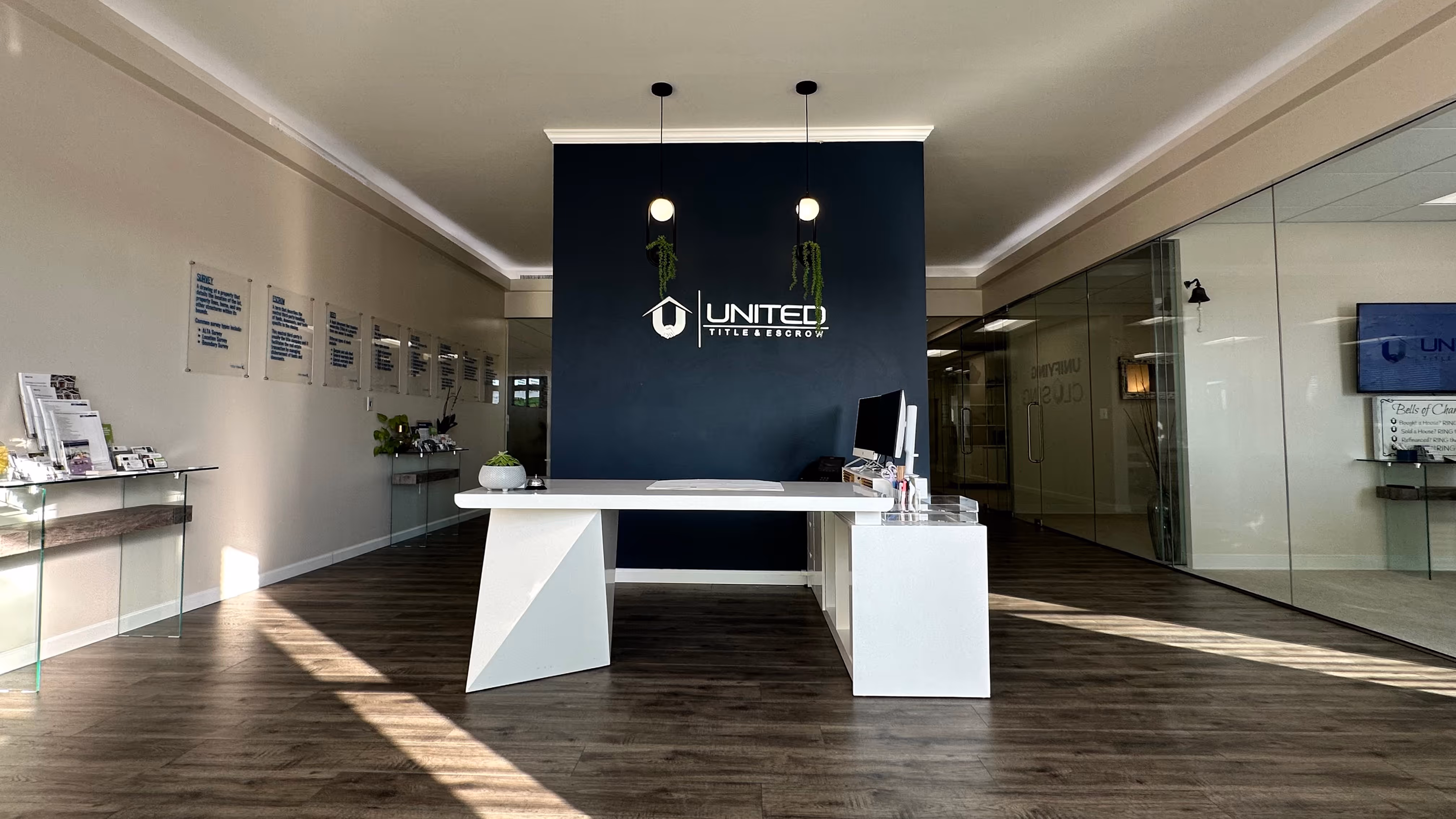 Modern office lobby with white desk, computer, hanging plants, and United Title & Escrow logo on dark accent wall.