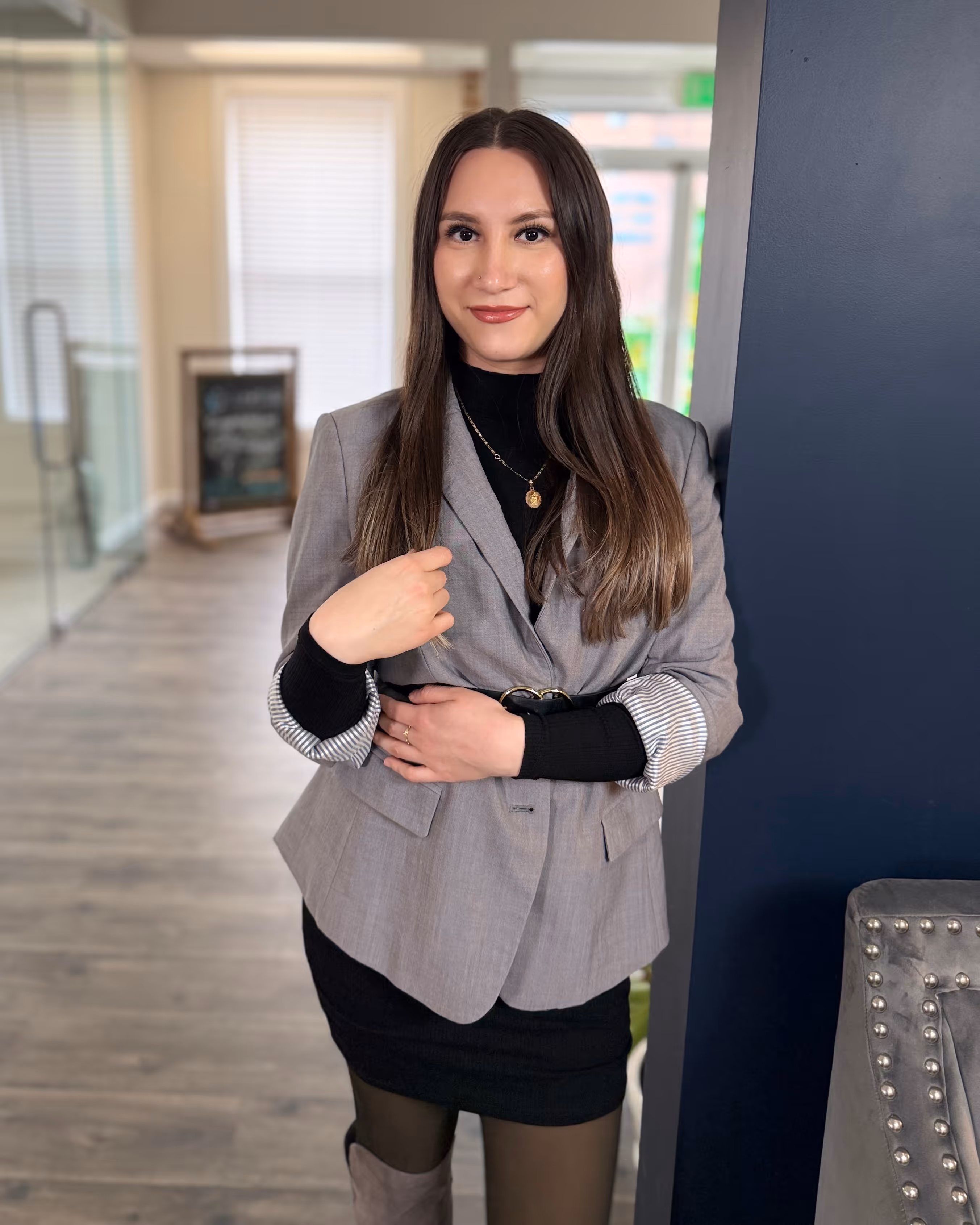 Woman with long brown hair wearing a gray blazer over a black dress, standing indoors against a dark blue wall.
