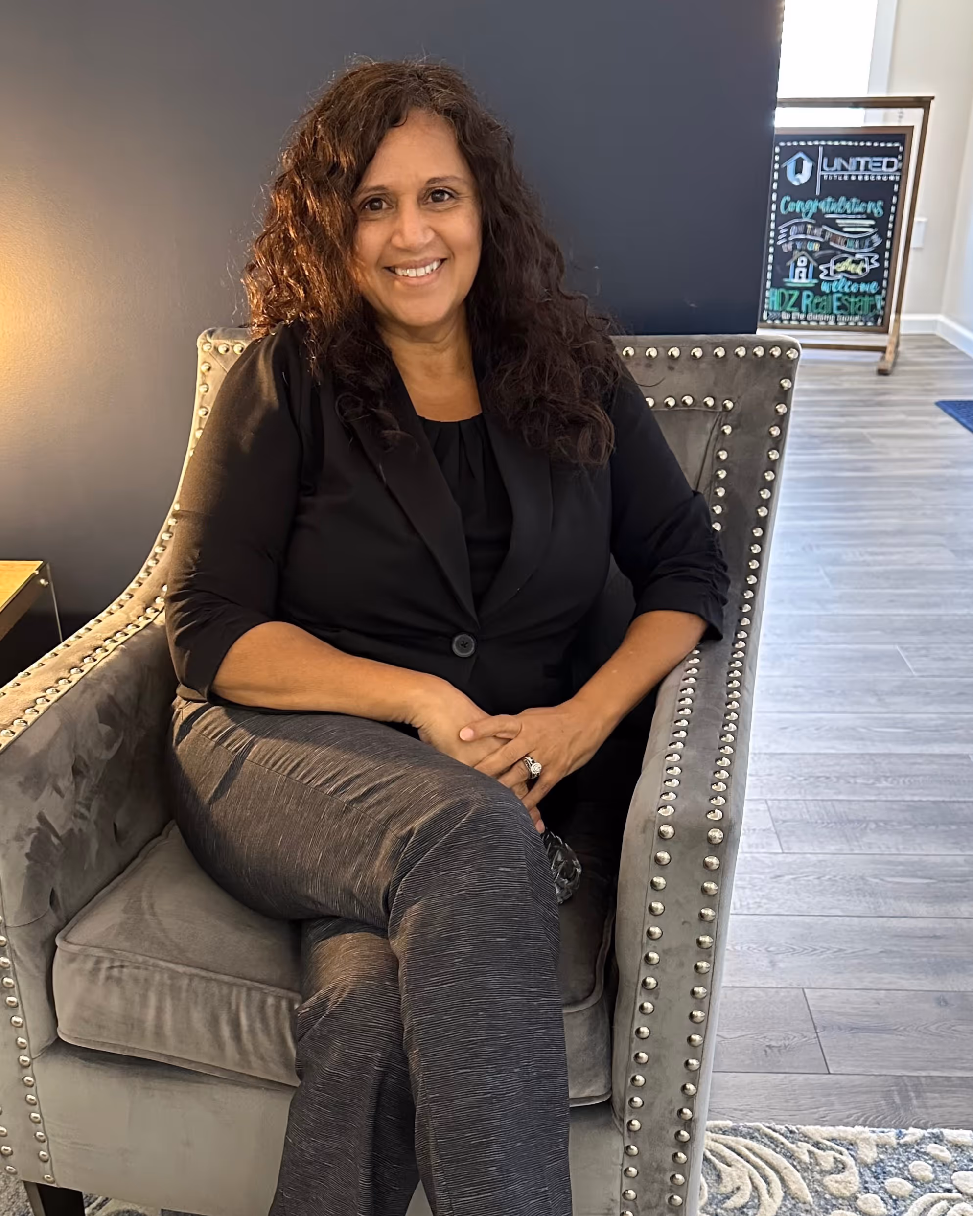 Smiling woman with curly hair sitting cross-legged in a gray studded armchair in a modern office setting.
