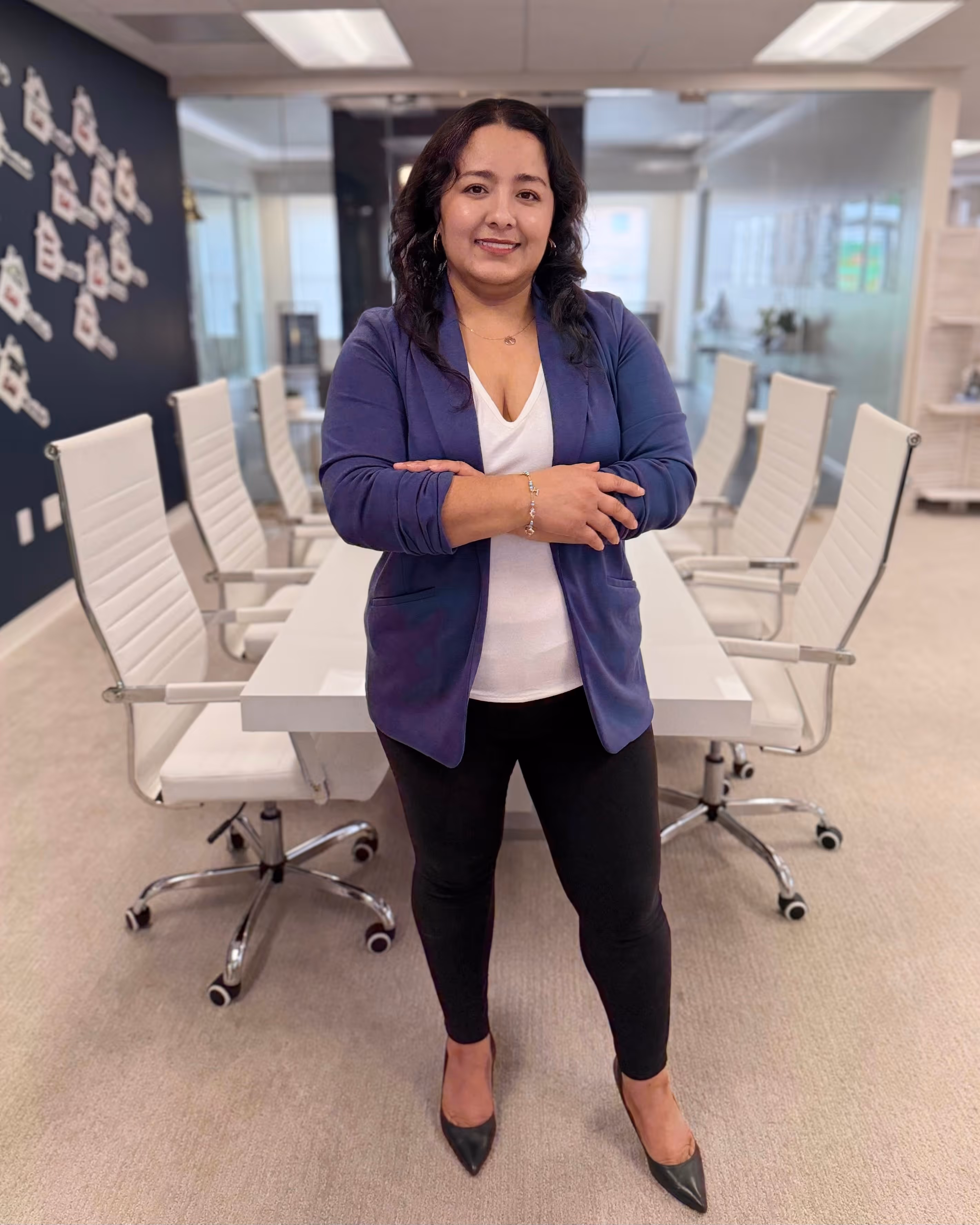 Woman with dark wavy hair wearing a blue blazer, white top, and black pants standing with arms crossed in a modern office conference room.