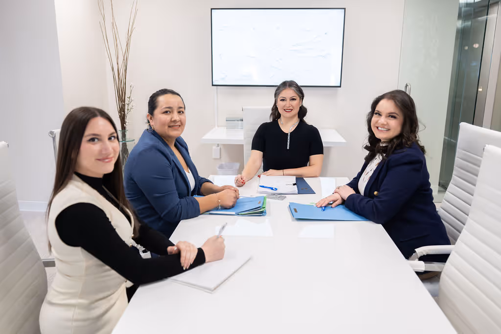 Four professional women seated around a white conference table, smiling and holding pens with folders and documents in front of them in a modern office.