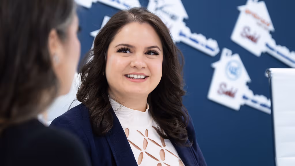 Smiling woman with dark hair talking to another person in a professional setting with a blue wall decorated with house-shaped sold signs.