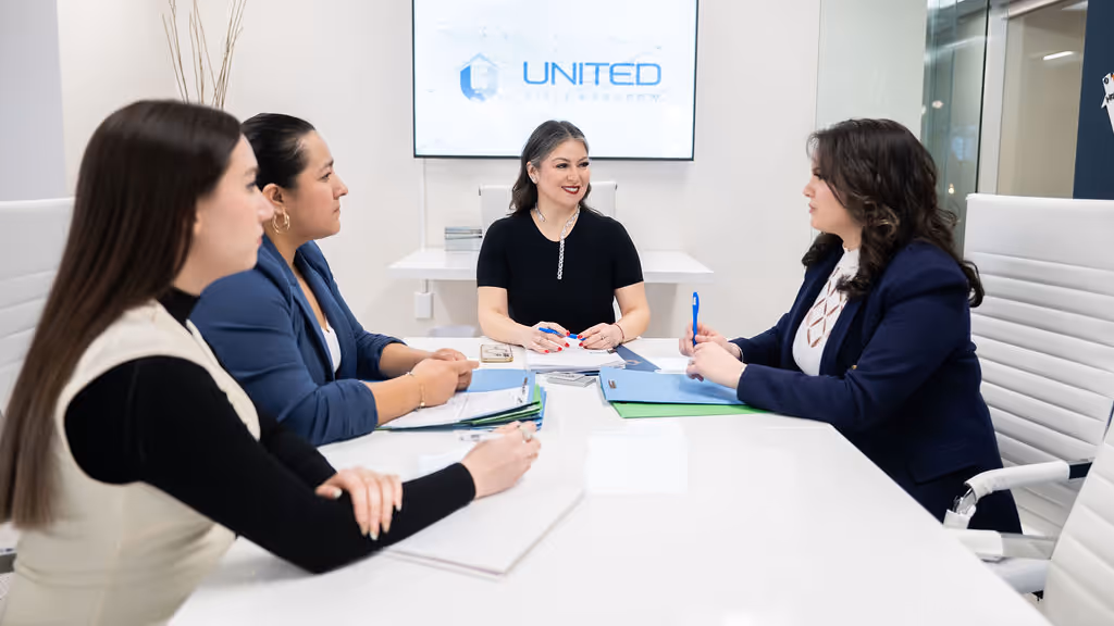 Four professional women seated around a white conference table engaged in a business meeting with a screen displaying United Technology logo behind them.