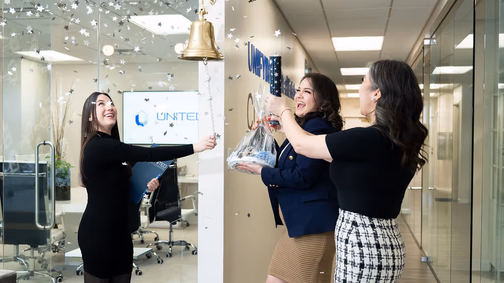 Three women celebrating in an office hallway as one rings a hanging bell with star confetti falling around them.