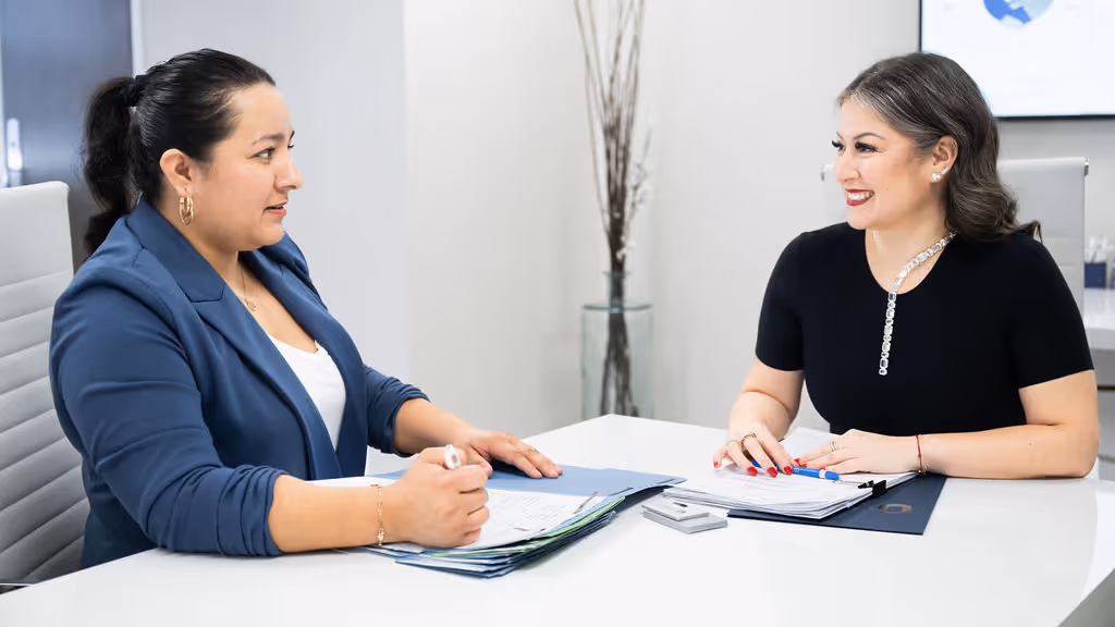 Two professional women sitting across a white table, discussing documents in an office setting.