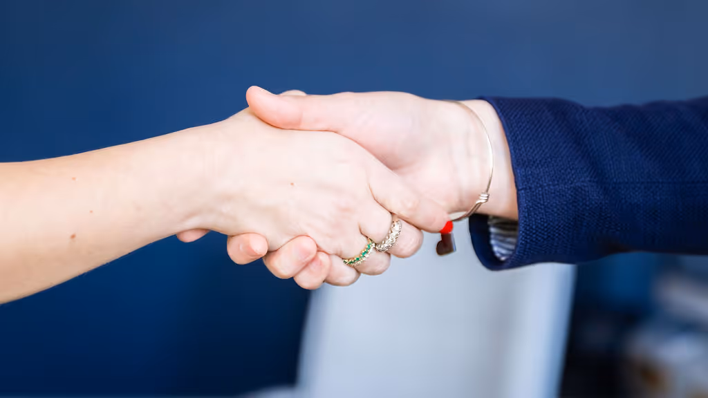 Two people shaking hands, one wearing a dark blue sleeve and rings, against a blue background.