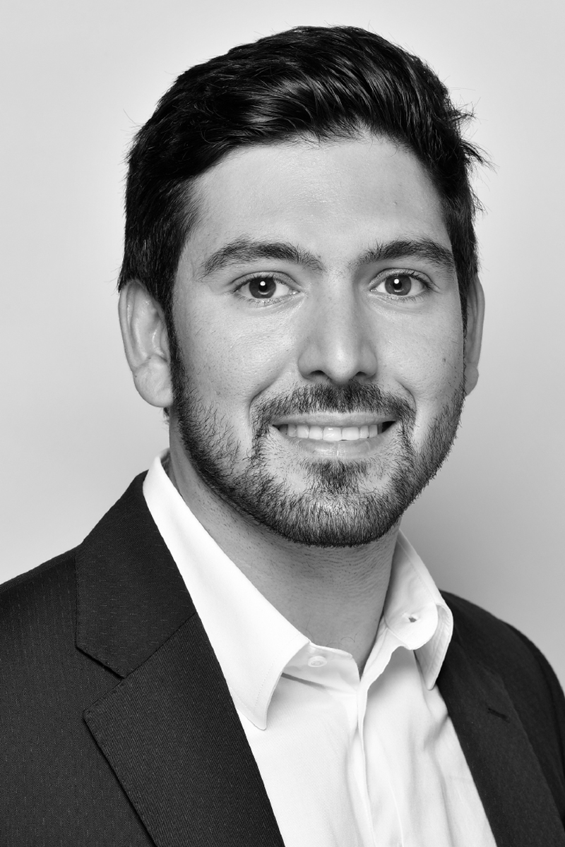 High-contrast black and white portrait of a man with slicked-back hair wearing a dark shirt against a dark background.