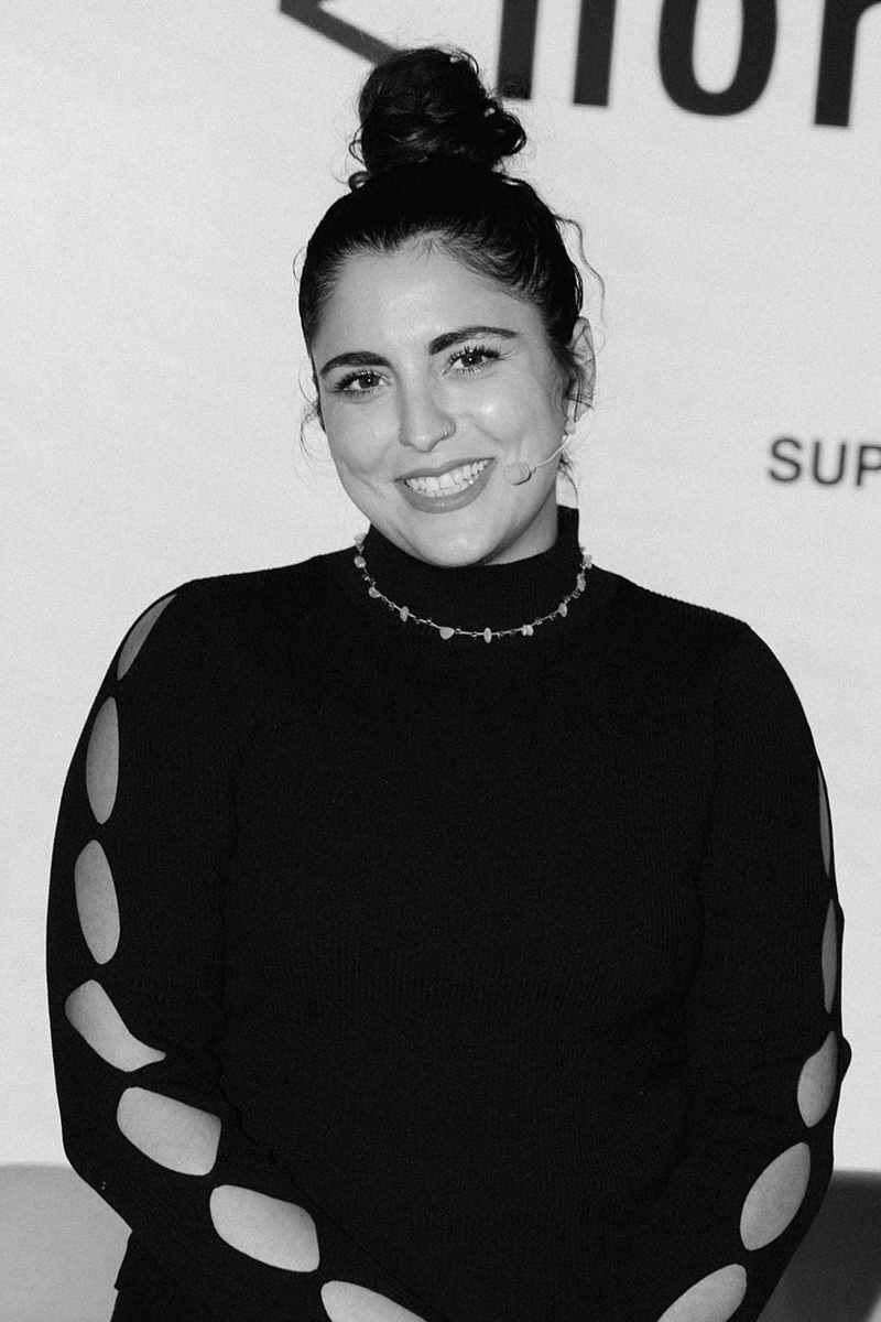 High-contrast black and white portrait of a man with slicked-back hair wearing a dark shirt against a dark background.