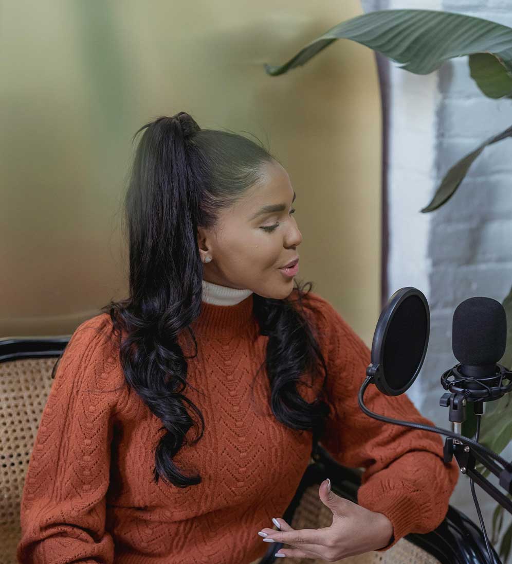 Woman in an orange sweater speaking into a microphone with a pop filter, seated in a chair beside a large plant.