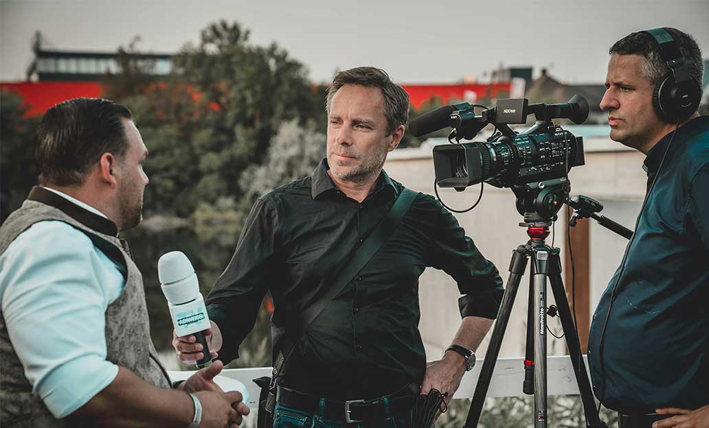 An outdoor interview scene with a man holding a microphone, another man operating a video camera on a tripod, and a cameraman wearing headphones.